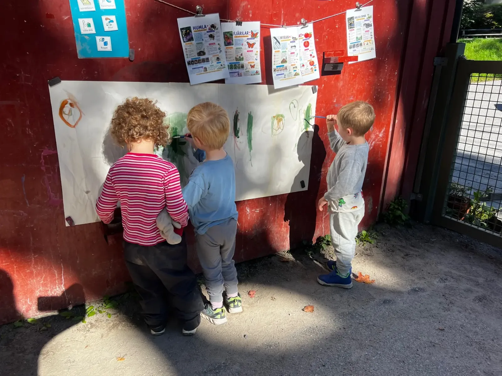Barn målar på stort papper utmed förskolans röda vägg